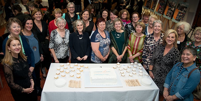A group photo of 25 current and former volunteers standing around a cake celebrating 15 years of Counterpart.