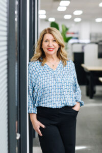 Smiling woman with light brown shoulder length hair and wearing a blue and white shirt leaning against a wall