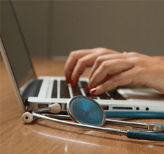 Close up of fingers typing on a laptop, with a stethoscope in the foreground.
