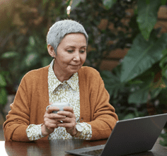 Relaxed woman with short grey hair sitting at an outdoor table drinking a coffee while she watches her laptop.