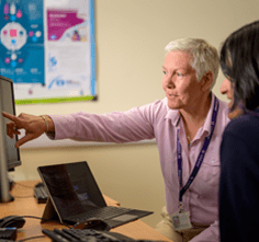 A casually dressed researcher with  lanyard around her neck points to a computer screen as she consults with a young woman, possibly a patient.