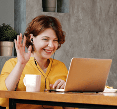 A woman with headphones on sits in front of her lap top and smiles and waves to someone she is meeting online.