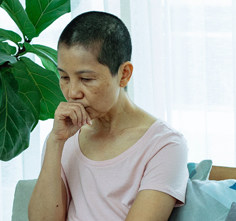 A woman with her hair shaved short sits in front of a leafy tropical houseplant. She is resting one hand against her chin and looks thoughtful.