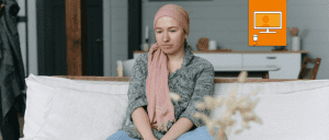 woman in headscarf sitting on couch staring into space. Online workshop icon with a computer in an orange square is in the top right hand corner of image.