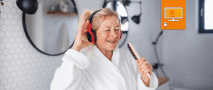 woman wearing a robe and headphones sings into hair brush. Our online workshop icon featuring a computer in orange box is in the top right hand corner.