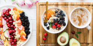 a bowl of fruits on bench to the left, bowl of fruit and yoghurt with cut avocado and a hot beveridge on a wooden chopping board to the right.