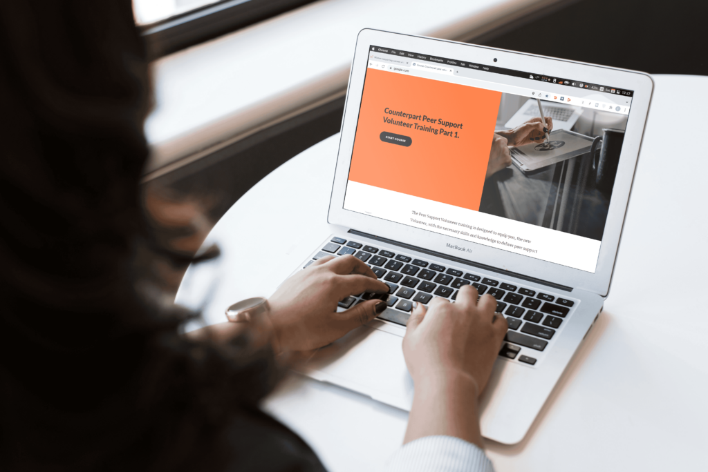 A woman using Counterpart's online volunteer training module on her laptop. On her screen is an orange block with text that says Counterpart Peer Support Volunteer Training part 1.