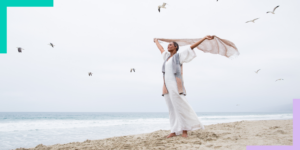 woman standing on beach with arms in the air holding a scarf that is flowing in the wind.