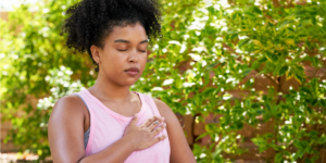 woman sitting with eyes closed and hand on chest deep breathing outside with trees in the background.