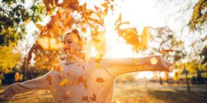 Women with outstretched arms throws up autumn leaves with the sun setting behind her.