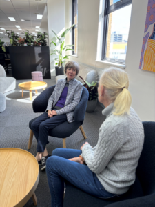 Two women sit on arm chairs having a casual chat in a quiet, airy office.