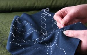Hands stitching light blue thread into navy blue fabric