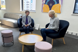 Two women sit on arm chairs having a casual chat in a quiet, airy office.