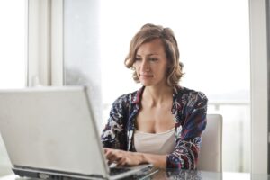 A woman sitting at a desk typing on a laptop, with a thoughtful expression on her face.