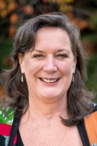 Portrait of a middle-aged woman with dark shoulder-length hair, fair skin and a warn smile. She is outside, with greenery in the background.