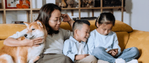 A woman and two school-aged children sit on a couch, one child is watching a phone, the other laughs, as the woman looks at them and smiles as she pats a dog sitting next to her.