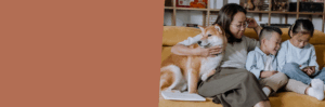 Image of mother, two children and their family dog sitting on the couch looking at a device and smiling.