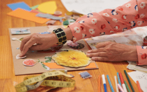 womans hand on collage piece sticking down images of flowers. A measuring tape and pencils sit in the foreground.